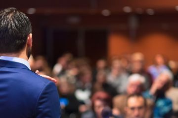 Man in a blue suit giving a presentation to a large, blurred audience in a conference room.